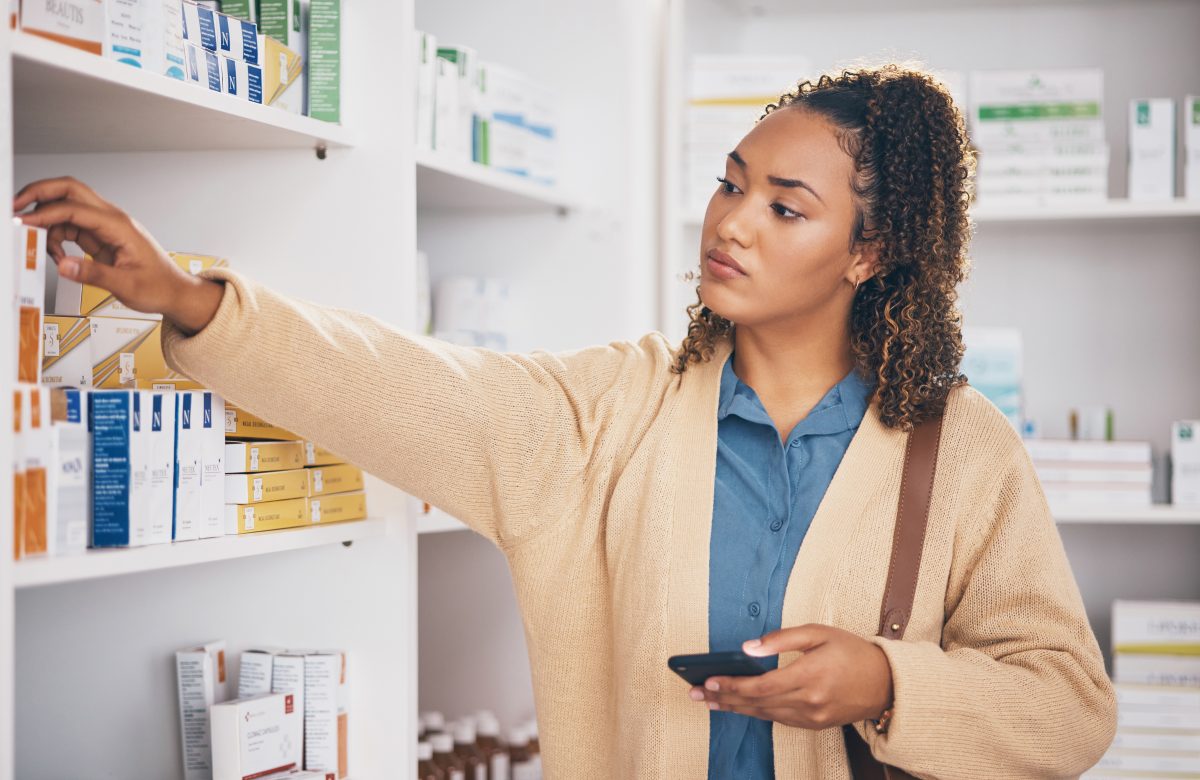 woman in a pharmacy picking up medicine while holding mobile phone in other hand